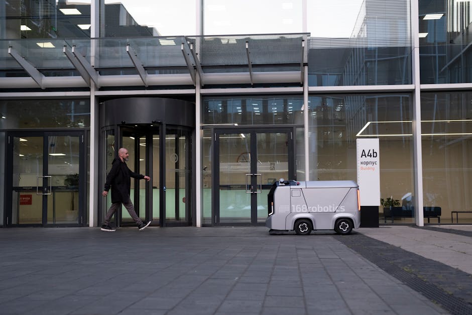 A gray 168 Robotics autonomous delivery robot on a paved path outside a modern building. A man walks past, showcasing AI logistics.