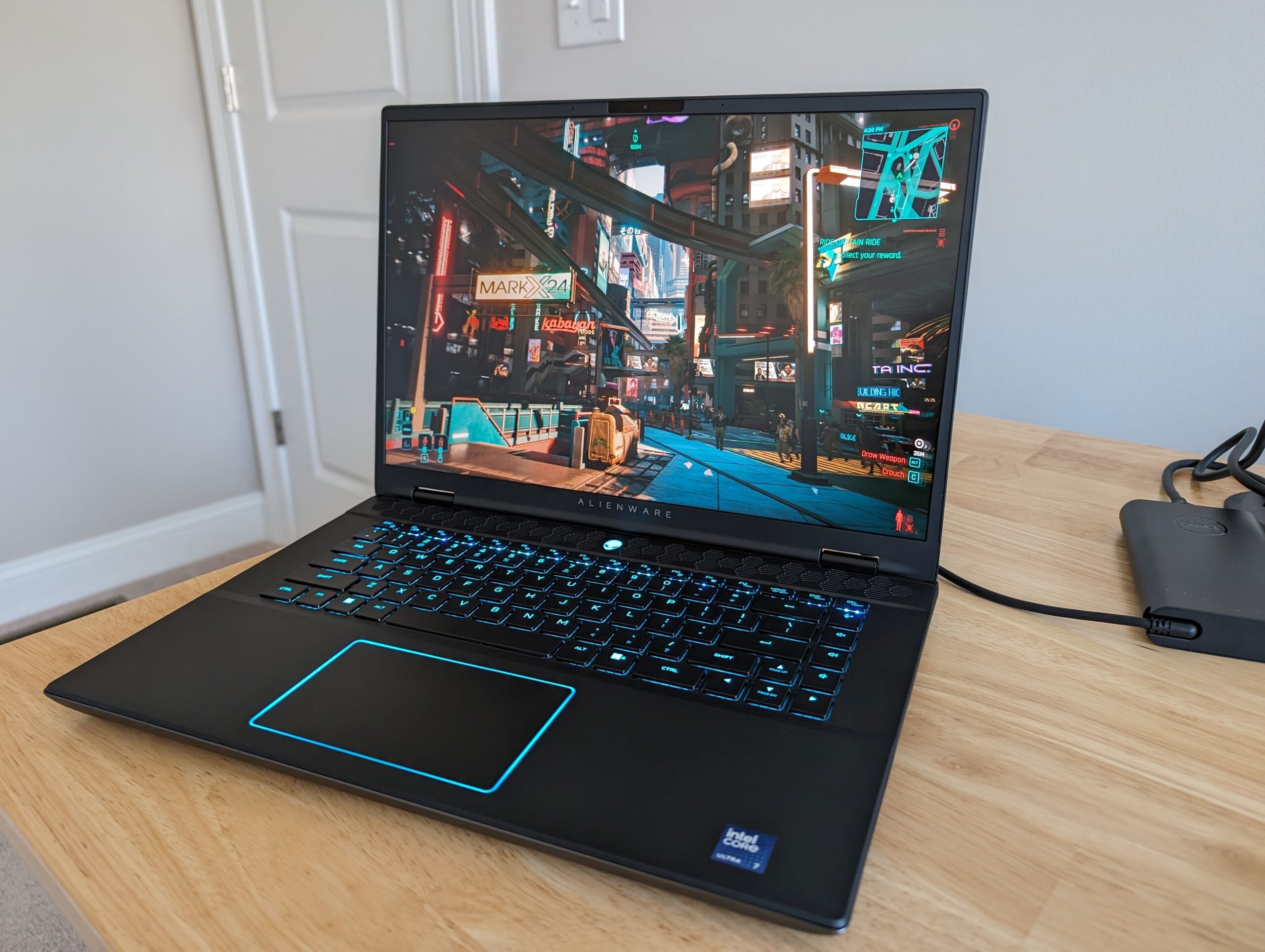 Alienware m16 R2 gaming laptop open on a wooden desk, displaying a vibrant game with a bright blue backlit keyboard and touchpad.