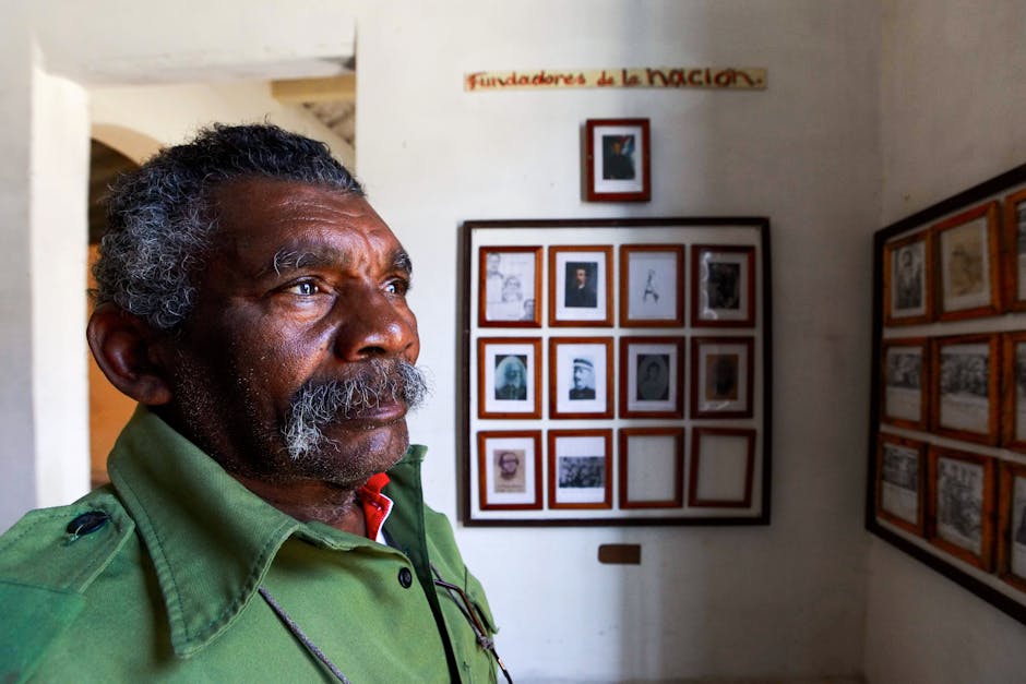 Mature man with gray mustache and green shirt looking at a wall of framed portraits, 'Fundadores de la Nacion' sign.