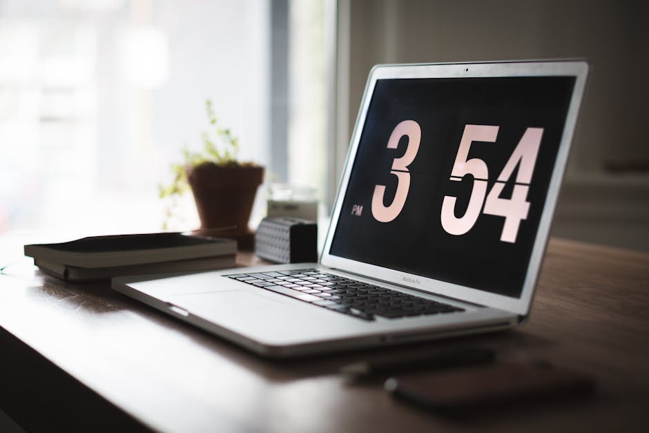 Silver laptop on a wooden desk displaying a digital clock (3:54 PM), with books and a plant in a focused solopreneur workspace.