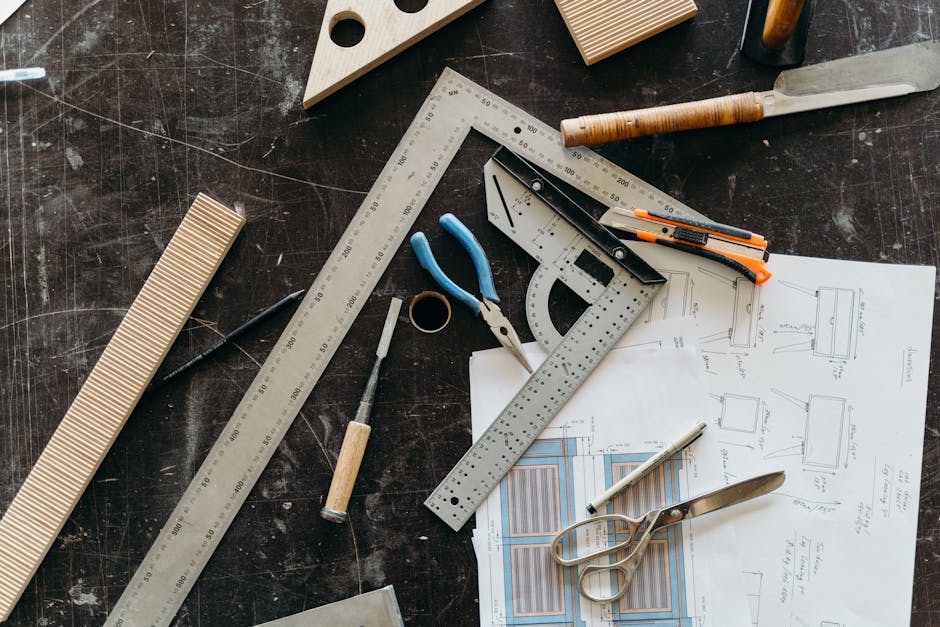 Overhead view of a design workbench with rulers, pliers, scissors, utility knife, and paper blueprints with sketches.